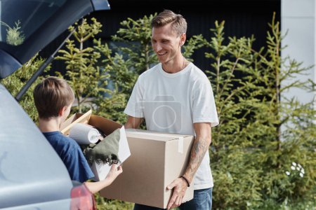 Téléchargez les photos : Portrait de père et fils souriants portant des boîtes à la voiture tout en se déplaçant dans une nouvelle maison éclairée par la lumière du soleil à l'extérieur - en image libre de droit