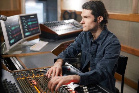 Téléchargez les photos : Portrait de producteur de musique masculin utilisant une station de travail audio numérique tout en travaillant avec des artistes dans un studio d'enregistrement professionnel - en image libre de droit