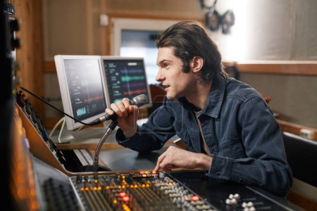 Téléchargez les photos : Portrait latéral de jeune homme parlant au microphone au poste de travail audio dans un studio d'enregistrement, industrie de la production musicale - en image libre de droit