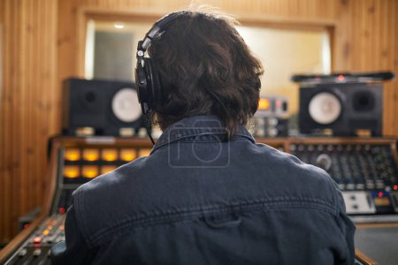 Photo for Back view of man wearing headphones at audio workstation in professional recording studio, copy space - Royalty Free Image