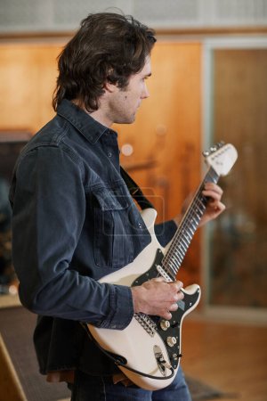 Photo for Vertical portrait of handsome young man playing electric guitar in recording studio and composing new music - Royalty Free Image