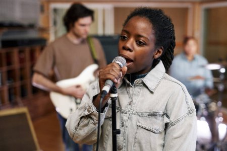 Photo for Waist up portrait of young black woman singing to microphone while recording music with band in professional studio - Royalty Free Image
