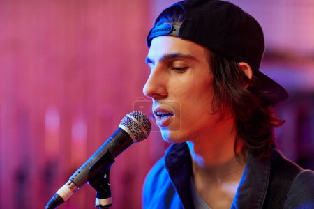 Photo for Profile portrait of young man singing to microphone in colored studio lights, copy space - Royalty Free Image