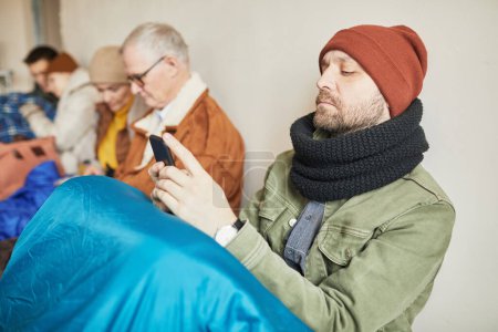 Photo for Side view portrait of bearded Caucasian man hiding in refugee shelter and holding smartphone, copy space - Royalty Free Image