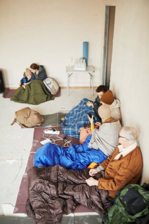 Foto de Vista de ángulo alto en el grupo de refugiados en el refugio del campamento sentado en alfombras con sacos de dormir - Imagen libre de derechos