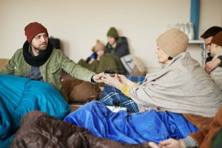 Téléchargez les photos : Groupe de réfugiés caucasiens partageant une tasse de thé tout en se cachant dans un abri ensemble pendant la guerre ou la crise couverte de couvertures - en image libre de droit