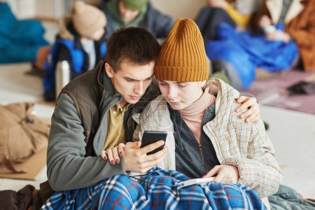 Photo for Front view portrait of young Caucasian couple hiding in refugee shelter covered with blanket on floor and holding smartphone - Royalty Free Image