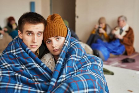 Téléchargez les photos : Vue de face portrait de jeune couple caucasien caché dans un refuge pour réfugiés recouvert d'une couverture sur le sol et regardant la caméra, espace de copie - en image libre de droit