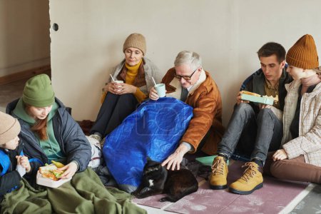 Photo for Group of Caucasian refugees eating food while hiding in shelter on floor covered with blankets - Royalty Free Image