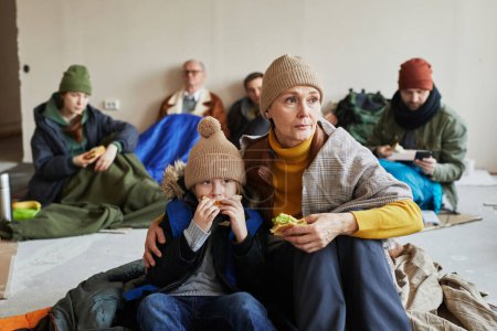 Bedrängte kaukasische Flüchtlingsfamilie isst Sandwiches und versteckt sich in einer Unterkunft auf dem Boden, der mit Decken bedeckt ist
