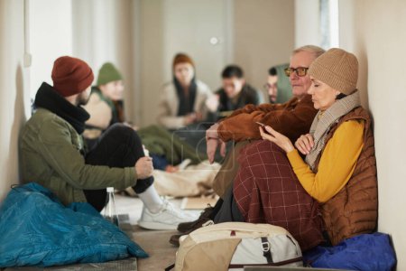 Téléchargez les photos : Portrait latéral de réfugiés caucasiens assis sur le sol dans un abri couvert de couvertures - en image libre de droit