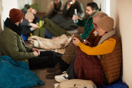 Téléchargez les photos : Portrait latéral de réfugiés âgés se cachant dans un abri pendant la guerre ou la crise et tenant un smartphone - en image libre de droit