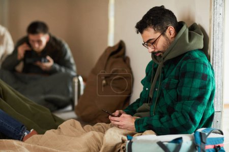 Photo for Side view portrait of young man hiding in shelter during war or crisis and holding smartphone - Royalty Free Image