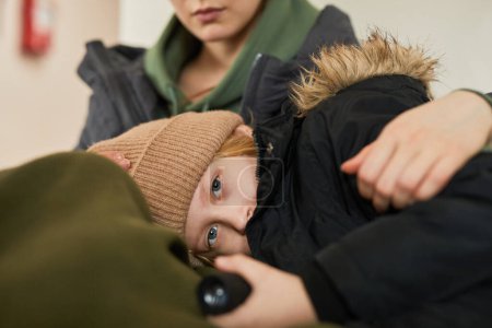 Téléchargez les photos : Portrait d'enfant blond caucasien allongé dans les mères genoux à l'abri des réfugiés et regardant la caméra avec les yeux bleus - en image libre de droit