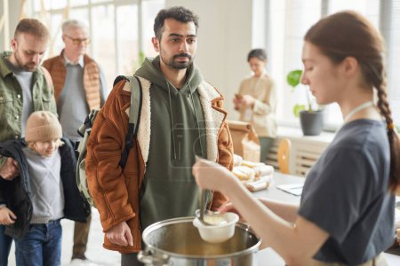 Photo for Portrait of young man standing in line at soup kitchen with young woman giving out simple meals to people in need - Royalty Free Image