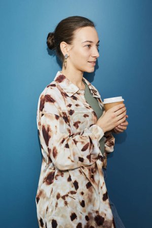 Photo for Minimal side view portrait of young businesswoman holding coffee cup against blue wall, shot with flash - Royalty Free Image