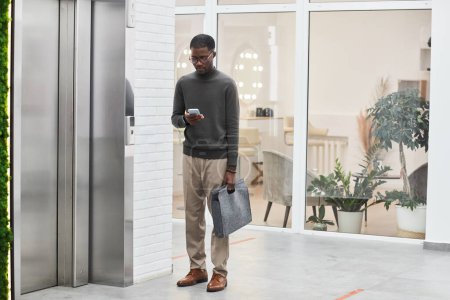 Foto de Retrato mínimo de larga duración del joven hombre de negocios negro de pie junto al ascensor en el moderno edificio de oficinas, espacio para copiar - Imagen libre de derechos