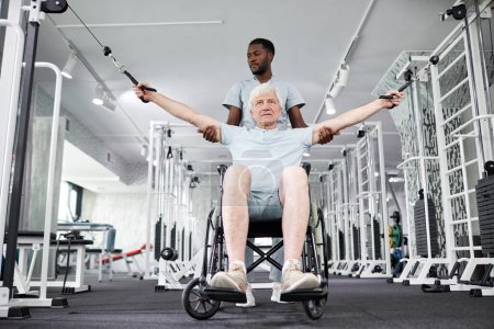 Foto de Retrato de bajo ángulo del terapeuta de rehabilitación afroamericano que ayuda a un hombre mayor en silla de ruedas a hacer ejercicios físicos en el gimnasio - Imagen libre de derechos