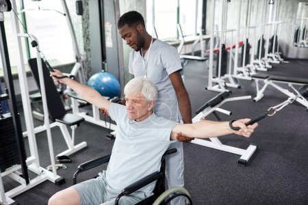 Foto de Retrato de un joven terapeuta de rehabilitación asistiendo a un hombre mayor en silla de ruedas con ejercicios físicos en el gimnasio - Imagen libre de derechos