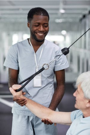 Téléchargez les photos : Portrait vertical d'un thérapeute de réadaptation souriant assistant un aîné au gymnase de la clinique - en image libre de droit
