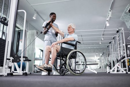 Photo for Low angle portrait of senior man using wheelchair in gym with rehabilitation therapist assisting, copy space - Royalty Free Image