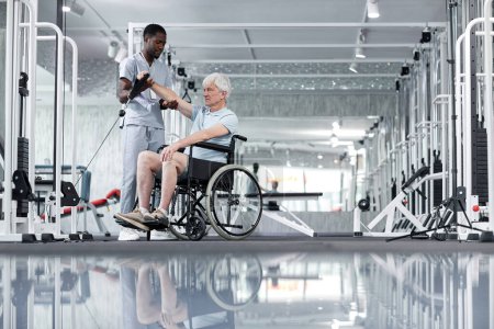 Foto de Amplia vista del ángulo en el hombre mayor de pelo blanco usando silla de ruedas en el gimnasio con la asistencia del terapeuta de rehabilitación, espacio de copia - Imagen libre de derechos