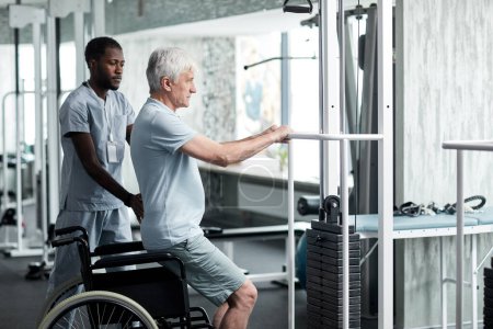 Photo for Side view portrait of senior man doing rehabilitation exercises in gym at medical clinic with therapist assisting - Royalty Free Image