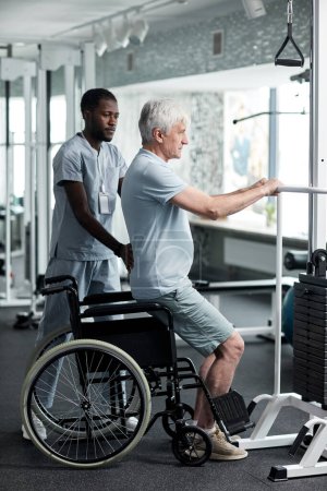 Photo for Full length portrait of senior man doing rehabilitation exercises in gym at medical clinic with therapist assisting - Royalty Free Image