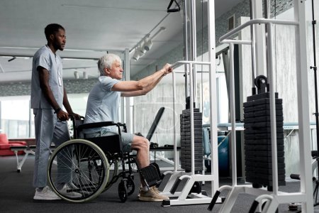 Photo for Side view portrait of senior man in wheelchair using exercise machines at rehabilitation clinic, copy space - Royalty Free Image