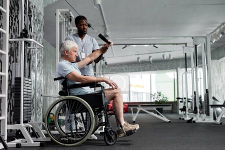 Foto de Retrato de vista lateral del hombre mayor usando silla de ruedas en el gimnasio y haciendo ejercicios de rehabilitación con asistente, espacio para copiar - Imagen libre de derechos