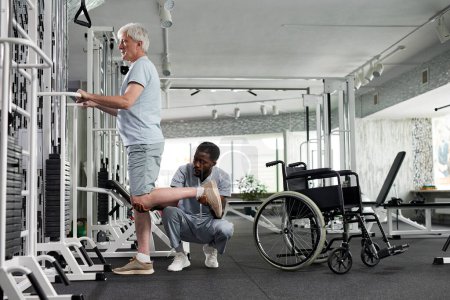 Photo for Side view of senior man doing rehabilitation exercises in gym with therapist assisting, copy space - Royalty Free Image