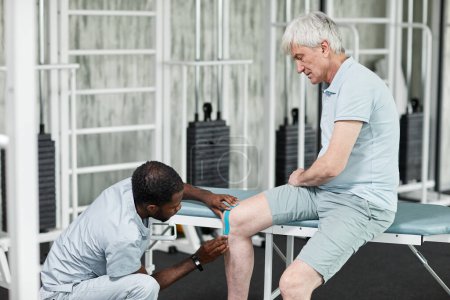 Photo for Portrait of rehabilitation specialist putting taping at knee of senior patient in physiotherapy clinic, copy space - Royalty Free Image