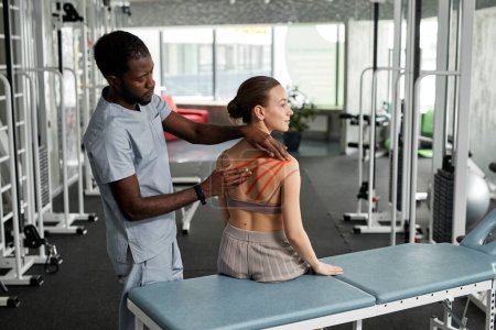 Téléchargez les photos : Portrait du thérapeute de réadaptation mettant la bande de kinesio sur le dos et l'épaule de la jeune femme dans la clinique de physiothérapie, espace de copie - en image libre de droit