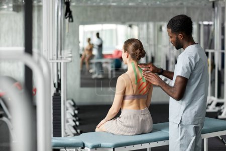 Téléchargez les photos : Thérapeute de réadaptation mettant la bande de kinesio sur le dos et l'épaule de la jeune femme en séance de physiothérapie - en image libre de droit
