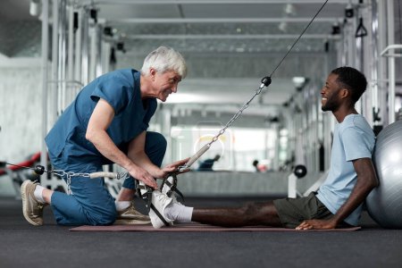 Photo for Portrait of smiling senior therapist helping young man with rehabilitation exercises in gym, copy space - Royalty Free Image