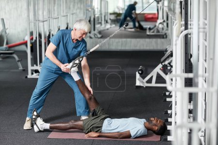 Téléchargez les photos : Portrait latéral de thérapeute senior souriant aidant jeune homme avec des exercices de réadaptation dans la salle de gym, espace de copie - en image libre de droit