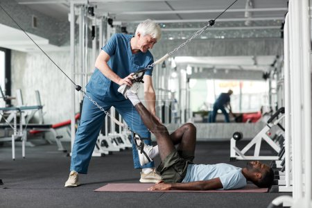 Photo for Side view at physiotherapy specialist helping young man with rehabilitation exercises in gym, copy space - Royalty Free Image