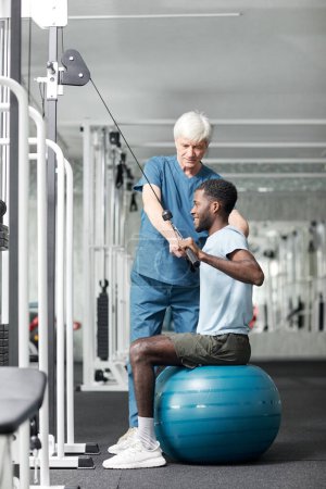 Téléchargez les photos : Portrait complet d'un spécialiste de physiothérapie aidant un jeune homme afro-américain à faire des exercices de réadaptation dans une salle de gym, un espace de copie - en image libre de droit