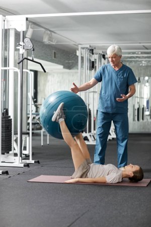 Photo for Vertical portrait of young woman doing rehabilitation exercises on floor at physiotherapy clinic with medic assisting - Royalty Free Image