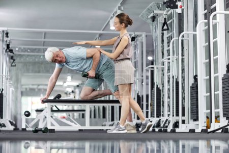 Téléchargez les photos : Portrait complet de l'homme âgé faisant des exercices sur banc à la salle de gym avec entraîneur de conditionnement physique féminin aider, copier l'espace - en image libre de droit