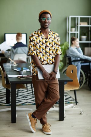 Téléchargez les photos : Portrait complet de jeune homme noir portant un motif jaune vif et regardant la caméra tout en restant au bureau - en image libre de droit