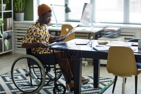 Téléchargez les photos : Portrait complet de jeune homme noir en fauteuil roulant portant de façon créative tout en travaillant dans le bureau, espace de copie - en image libre de droit