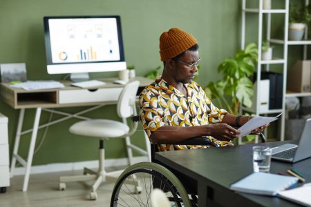 Foto de Retrato de un joven negro en silla de ruedas con estilo creativo mientras trabaja en la oficina, espacio para copiar - Imagen libre de derechos