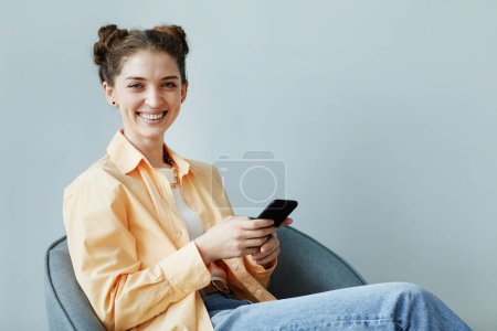 Foto de Retrato mínimo de una joven despreocupada usando un teléfono inteligente mientras está sentada contra la pared azul en la oficina y sonriendo a la cámara, espacio para copiar - Imagen libre de derechos
