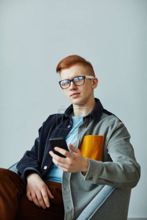Foto de Retrato mínimo de un joven creativo usando un teléfono inteligente mientras se sienta contra la pared azul en la oficina y sonríe a la cámara - Imagen libre de derechos