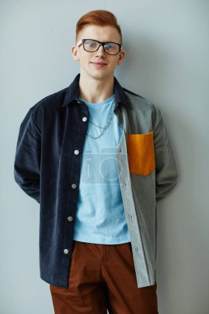 Téléchargez les photos : Portrait minimal de jeune homme créatif portant des lunettes tout en se tenant contre le mur bleu et souriant à la caméra - en image libre de droit