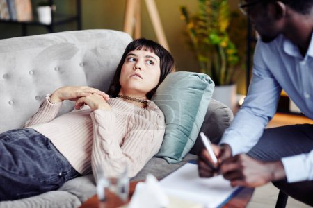 Foto de Mujer joven acostada en un sofá en una sesión de terapia, confesando problemas y mirando a su psicoterapeuta - Imagen libre de derechos