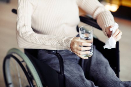 Photo for Young woman in wheelchair drinking water and wiping tears with napkin after emotional therapy session with psychologist - Royalty Free Image