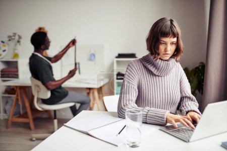 Téléchargez les photos : Concentré jeune femme travaillant sur ordinateur portable dans un bureau créatif avec son collègue - en image libre de droit