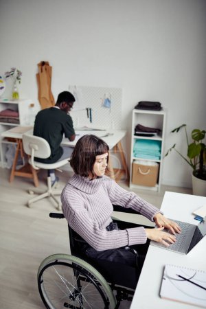 Foto de Joven empresaria con discapacidad que trabaja en el portátil en el estudio de moda, su colega de costura en segundo plano - Imagen libre de derechos